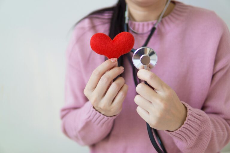 Unrecognizable female in pink sweater with stethoscope on neck standing on white background with red heart in hand in daylight